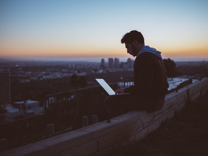 Photograph of a man sat on a wall using a laptop in the evening